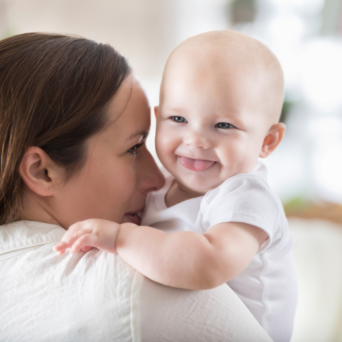 Why Your Baby or Toddler Isn’t Sleeping — Take the Free Sleep Quiz | Otterly Thriving Smiling mom holding a smiling baby with her tongue out on her shoulder in a baby sleep sack.